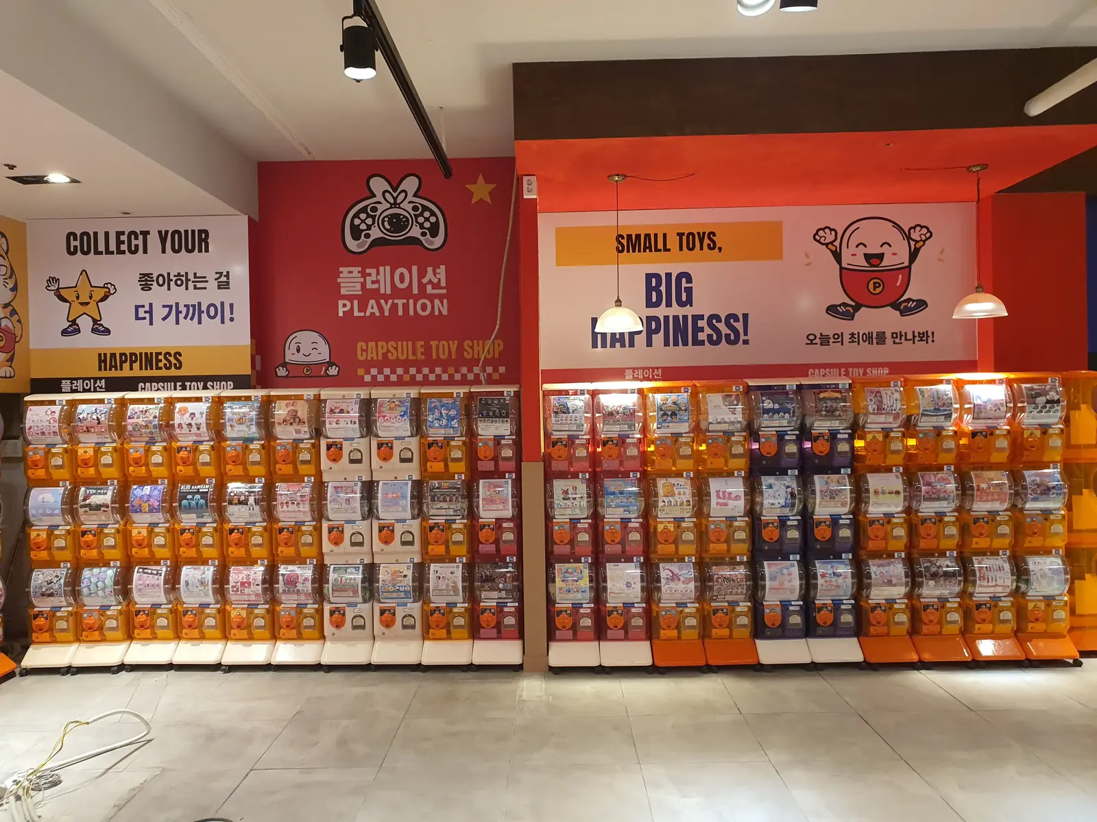 SMALL TOYS, BIG HAPPINESS 문구와 캡슐 캐릭터가 그려진 밝은 벽면 아래의 캡슐토이 기기들